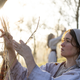 A woman in a white headscarf gathers sticks as sun peeks through the trees.